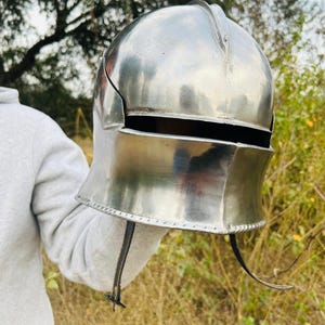 May include: A polished, silver-colored medieval helmet with a rounded top and a curved lower section. The helmet has a visor opening and a chin strap. The background is blurred, with green and brown tones.