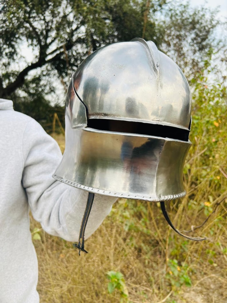 May include: A polished, silver-colored medieval helmet with a rounded top and a curved lower section. The helmet has a visor opening and a chin strap. The background is blurred, with green and brown tones.