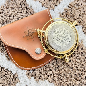 Op de afbeelding: Een rond navigatie-instrument van messing met een complexe wijzerplaat met Romeinse cijfers en concentrische cijferringen, bevestigd aan een goudkleurige ketting. Het wordt geleverd met een bruinleren etui met een kompasroos-opdruk.