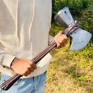 May include: A prop replica of a battle axe with a silver-colored head and a brown handle. The axe head has intricate engraved details. The handle is wrapped with a dark brown material and secured with a black band.