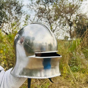 May include: A polished, silver-colored medieval helmet with a rounded top and a visor. The helmet has a curved neck guard and a leather strap with a buckle. The background features trees and foliage.