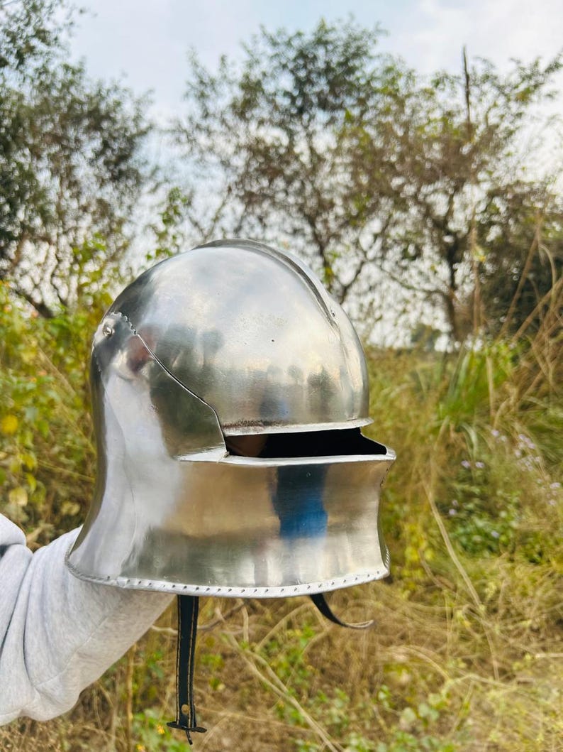 May include: A polished, silver-colored medieval helmet with a rounded top and a visor. The helmet has a curved neck guard and a leather strap with a buckle. The background features trees and foliage.