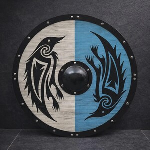 May include: A round wooden shield with a black rim and a central black boss. The shield is divided vertically, with one half white and the other light blue. Each half features a black stylized bird design.