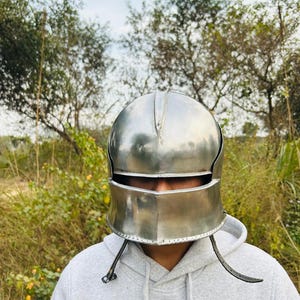 May include: A polished silver metal helmet with a visor and cheek guards. The helmet is worn over a gray hooded sweatshirt. The background features green foliage and a cloudy sky.