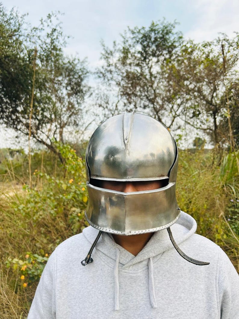 May include: A polished silver metal helmet with a visor and cheek guards. The helmet is worn over a gray hooded sweatshirt. The background features green foliage and a cloudy sky.