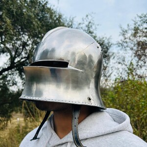 May include: A polished, silver-colored medieval helmet with a visor and leather chin straps. The helmet is designed to protect the head and face. The background features trees and a cloudy sky.