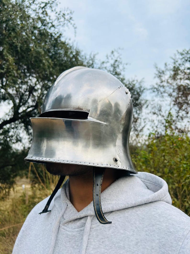 May include: A polished, silver-colored medieval helmet with a visor and leather chin straps. The helmet is designed to protect the head and face. The background features trees and a cloudy sky.