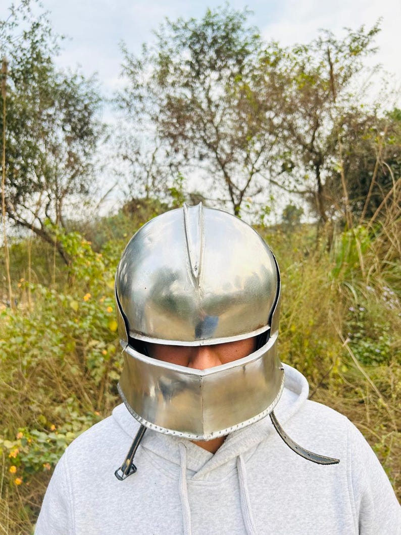 May include: A polished silver metal helmet with a visor and cheek guards. The helmet is designed with a rounded top and a defined brow line. The background features green foliage and trees.