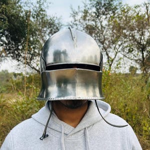 May include: A polished silver metal helmet with a rounded top and a horizontal eye slit. The helmet has a flared base and a vertical ridge on top. It is worn over a gray hooded sweatshirt.