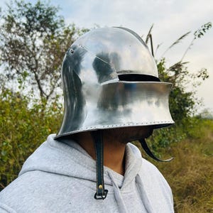 May include: A polished, silver-colored medieval helmet with a visor and leather chin straps. The helmet is designed to protect the head and face. The visor is down, obscuring the face. The helmet is made of metal and has a smooth, reflective surface.
