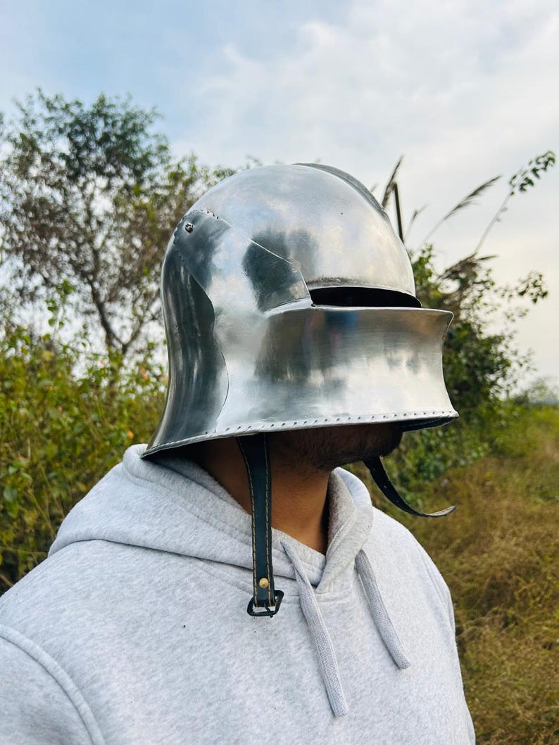 May include: A polished, silver-colored medieval helmet with a visor and leather chin straps. The helmet is designed to protect the head and face. The visor is down, obscuring the face. The helmet is made of metal and has a smooth, reflective surface.