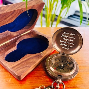 May include: A vintage-style compass with a quote engraved on the inside of the lid, presented in an open wooden box with blue velvet lining. The compass is made of brass and has a key chain attached.