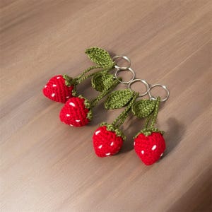 May include: Four handmade crochet strawberry keychains. Each keychain features a red strawberry with white seed details, green leaves, and a silver-colored keyring. The keychains are arranged on a wooden surface.