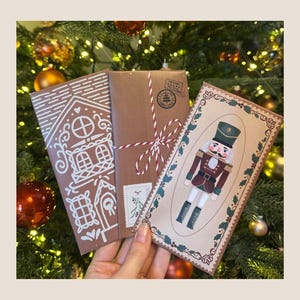 May include: Three Christmas-themed cards are held in front of a decorated Christmas tree. The cards feature a gingerbread house, a package tied with red and white twine, and a nutcracker illustration. The cards are brown, white, red, green, and black.