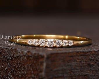 May include: A delicate gold ring with seven small, clear gemstones set in a row. The ring has a smooth, polished band and is displayed on a dark, textured surface. The text "Diamond Vows" is visible on the left side of the ring.