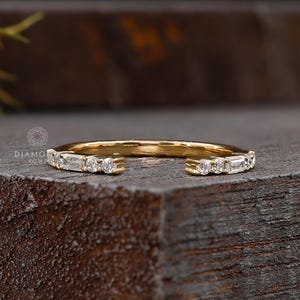 May include: A gold ring with an open design, set with small, clear gemstones. The ring features a mix of round and baguette-cut stones. The ring is displayed on a dark, textured wooden surface.