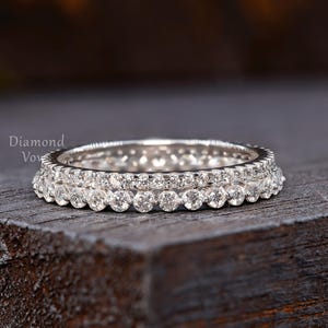 Op de afbeelding: Een zilverkleurige eeuwigheidsring met een doorlopende rij kleine, ronde, heldere stenen in een metalen zetting. De ring toont de tekst Diamond Vows aan de linkerkant. De ring heeft een gepolijste afwerking en een uniform, rond ontwerp.