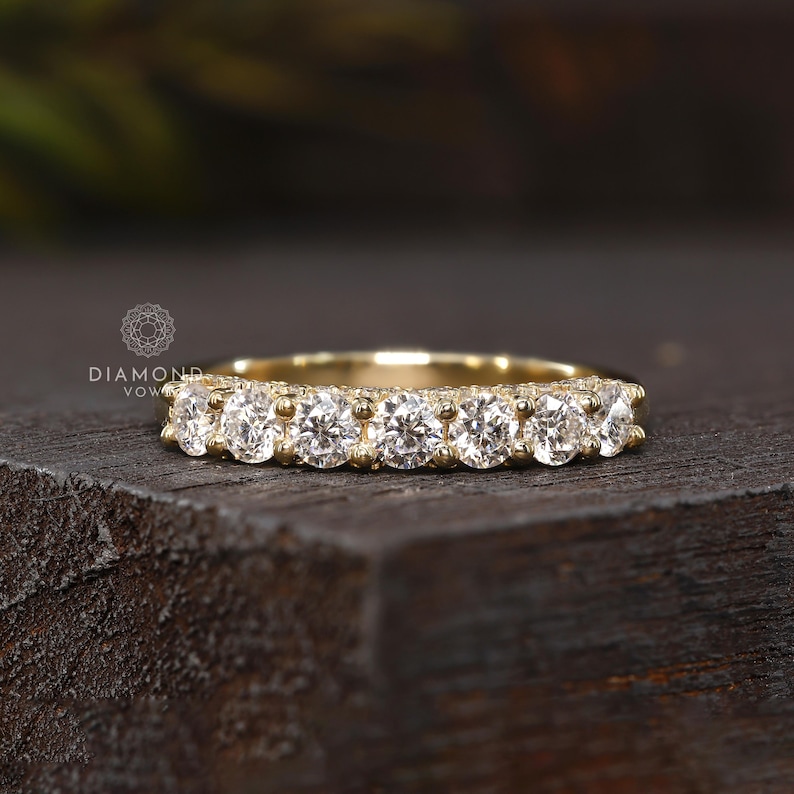 May include: A gold band ring featuring seven round-cut diamonds. The ring has a delicate design with small accent stones along the band. The ring is set on a dark wooden surface, showcasing the sparkle of the diamonds.