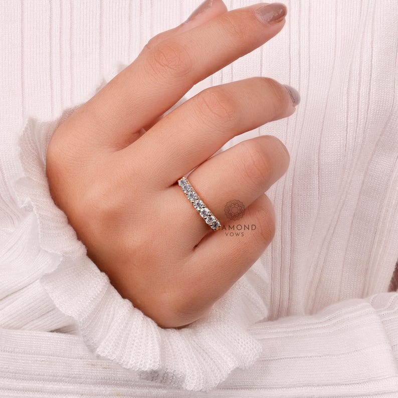May include: A gold band ring set with multiple round-cut diamonds. The ring is shown on a hand, with a white fabric background. The ring is a classic design, suitable for various occasions.