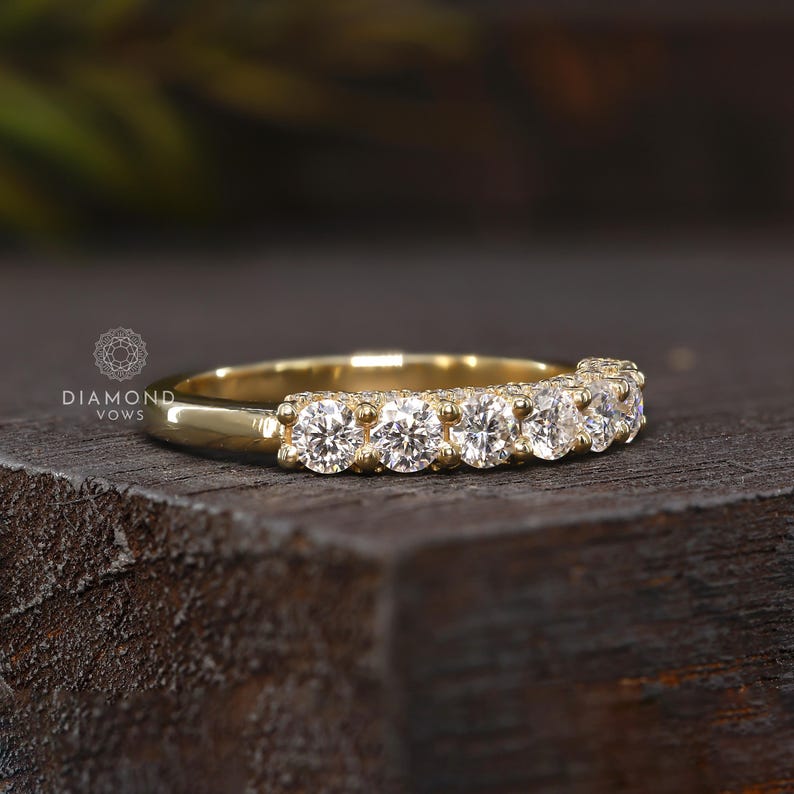 May include: A gold band ring featuring a row of six round-cut diamonds. The ring is set on a dark wooden surface. The brand name "DIAMOND VOWS" is visible in the image.