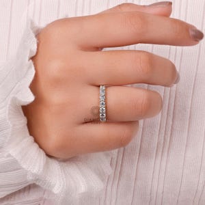 May include: A gold band ring with a row of clear, sparkling stones. The ring is on a person's finger, with a soft, neutral-colored background. The ring is a classic design, suitable for various occasions.