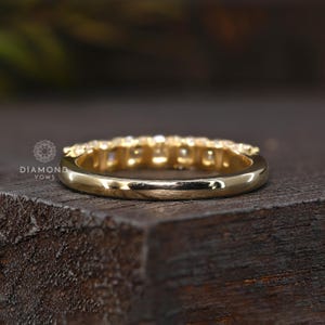 May include: A gold band ring with a row of small, clear gemstones set across the top. The ring is shown from a side angle, resting on a dark wooden surface. The ring is highly polished and reflects light.