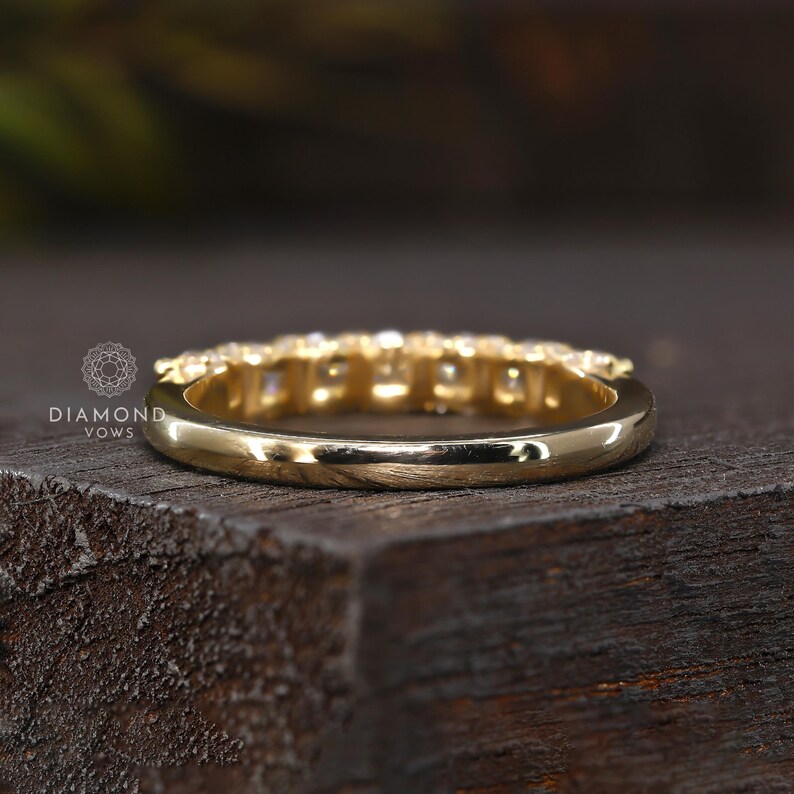 May include: A gold band ring with a row of small, clear gemstones set across the top. The ring is shown from a side angle, resting on a dark wooden surface. The ring is highly polished and reflects light.