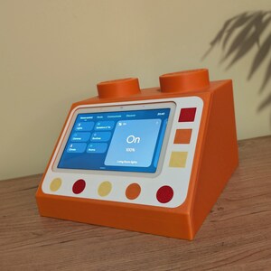 Könnte beinhalten: Ein orangefarbenes und weißes Smart-Home-Gerät in Form eines Bausteins, mit einem Tablet, das eine blaue Oberfläche anzeigt. Das Gerät hat zwei orangefarbene Zylinder oben und farbige Akzente. Der Bildschirm zeigt das Wort "On".