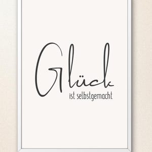 May include: A white framed wall art print featuring a cream-colored background with black text. The word "Glück" is written in a large, elegant script font, followed by the smaller, sans-serif text "ist selbstgemacht" centered below it.