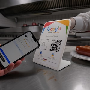 Könnte beinhalten: Ein weißer Google Reviews-Ständer mit einem QR-Code und dem Google-Logo, neben einem Smartphone, das eine Bewertungsseite anzeigt. Zwei Würstchen liegen auf einem Teller im Hintergrund.