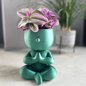 Könnte beinhalten: Türkisgrüner Meditationsfiguren-Pflanztopf mit einer Pflanze mit rosa und grünen Blättern. Die Figur befindet sich in einer sitzenden Yoga-Pose mit zusammengelegten Händen. Ein einzigartiger Dekorationsartikel.