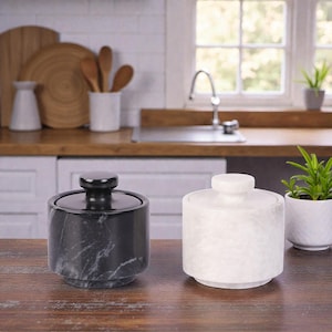 May include: Two marble canisters with lids. One is black with gray veining, the other is white. Both have a cylindrical shape and sit on a wooden surface. The background shows a kitchen setting with a window and plants.
