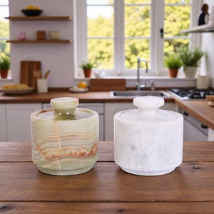 Pode incluir: Dois potes decorativos com tampa. Um é verde e castanho, o outro branco. Ambos são cilíndricos com base arredondada e um botão no topo. Estão sobre uma superfície de madeira numa cozinha.
