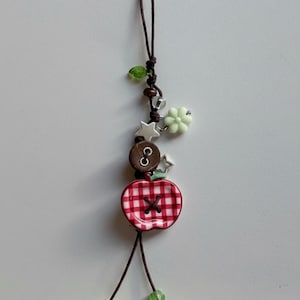 Puede incluir: Un colgante decorativo con un cordón marrón y varias cuentas, incluyendo un botón en forma de manzana con cuadros rojos y blancos, un botón circular marrón, una flor verde claro, estrellas plateadas y cuentas verdes y transparentes.