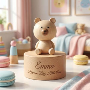 Pode incluir: Uma figura de urso de madeira está em cima de um cilindro de madeira, gravado com o nome "Emma" e a frase "Dream Big, Little One". O urso tem uma cabeça redonda, orelhas pequenas e um nariz castanho. A cena passa-se num quarto infantil com cores pastel.