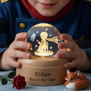 May include: A glowing snow globe featuring a seated figure with a scarf, surrounded by stars. The globe sits on a wooden base engraved with "Ethan Reach the Stars." A red rose and a fox figurine are also present.