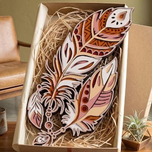 May include: Two decorative wooden feathers in a gift box. The feathers feature layered designs in white, peach, brown, and orange. The box is lined with straw and tied with a brown ribbon. The feathers are approximately 30 cm long.