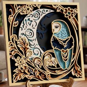 Op de afbeelding: Een meerlaags houten kunstwerk met een uil en een maan. De kleuren zijn blauw, bruin en crème, met bloemen- en bladaccenten. Het kunstwerk wordt gepresenteerd op een houten ezel.