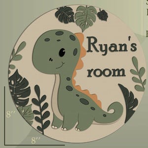 Op de afbeelding: Een ronde, lichtbeige plaat met een groene dinosaurusillustratie en de tekst "Ryan's room". De dinosaurus heeft oranje accenten en is omgeven door groene bladeren. De plaat heeft een diameter van 20 cm.