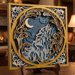 May include: A layered wooden art piece featuring a howling wolf silhouette against a night sky with a crescent moon and stars. The artwork is framed in gold and set on a wooden stand. The wolf is white and the background is blue.