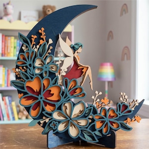 Può includere: Decorazione in legno a strati con una fata e una luna. La luna a mezzaluna blu scuro è adornata con fiori colorati nelle tonalità dell'arancione, del verde acqua e del crema. Una fata con un vestito rosso e ali color crema è seduta sulla luna.