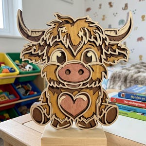 May include: A layered wooden sculpture of a cartoon Highland cow. The sculpture features a light brown and tan color scheme with a heart-shaped detail on the chest. The cow has large eyes and curved horns, sitting on a wooden base.
