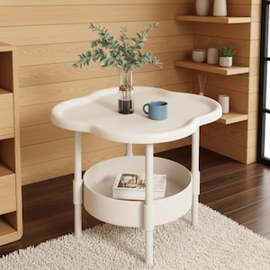 Pode incluir: Uma mesa lateral branca em forma de nuvem com uma prateleira inferior. Uma caneca azul e um vaso com vegetação estão em cima. A mesa tem uma prateleira inferior redonda e assenta em quatro pernas.