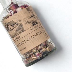 Puede incluir: Una botella de vidrio transparente llena de hierbas y flores secas. La botella tiene una etiqueta marrón con el texto "SAMOTNA CHATA" y un dibujo de una casa. La etiqueta también incluye el texto "MASA NETTO 60g".