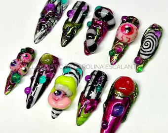Puede incluir: Un conjunto de diez uñas postizas con diseños 3D complejos, rayas blancas y negras, acentos metálicos en verde y rosa, y motivos de globos oculares incrustados. El texto CLAWISTIC BY CAROLINA ESCALANTE es visible en el centro del conjunto.