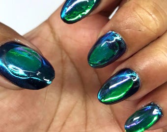 May include: Oval-shaped fingernails featuring a high-gloss, metallic chrome finish. The polish displays a vibrant color-shifting effect, transitioning between deep navy blue, emerald green, and subtle violet highlights across the nail surface.