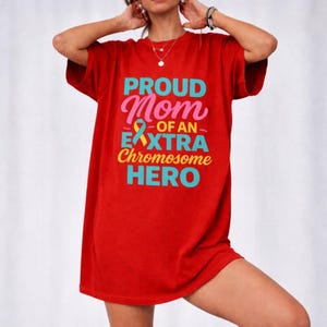 Op de afbeelding: Rood T-shirt met de tekst "PROUD Mom - OF AN - EXTRA Chromosome HERO" in teal, roze en geel. Het shirt heeft een ronde hals en korte mouwen. Het shirt heeft een effen rode kleur.