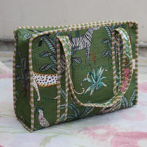May include: A rectangular quilted tote bag in olive green featuring a repeating print of zebras, leopards, and tropical plants. The bag has two matching fabric handles and is finished with a pink and white striped trim along all edges.