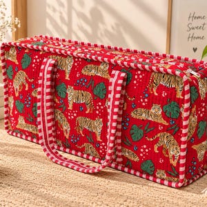 May include: A rectangular quilted fabric storage bag in a vibrant red color featuring a repeating pattern of tigers, green monstera leaves, and blue berries. The bag has two red and white striped fabric handles and a zippered top closure.