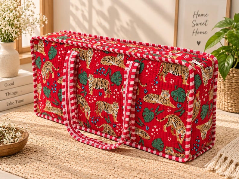 May include: A rectangular quilted fabric storage bag in a vibrant red color featuring a repeating pattern of tigers, green monstera leaves, and blue berries. The bag has two red and white striped fabric handles and a zippered top closure.